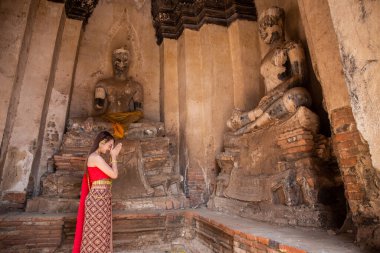 Geleneksel kırmızı Tayland elbisesi ve altın süslemeleri giymiş genç bir kadın Buda heykellerine tapınıyor ve büyük Buda heykellerini karşılıyor. Wat Chaiwatthanaram Ayutthaya