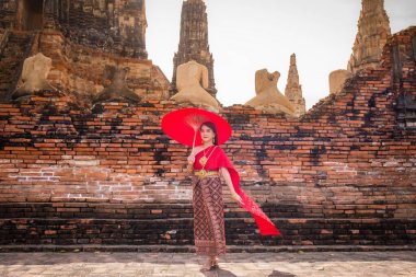 Geleneksel kırmızı Tayland elbisesi ve altın aksesuarları giyen genç bir kadın tarihi alanda geleneksel bir şemsiye tutuyor. Tayland ulusal kostümü