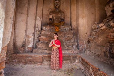 Geleneksel kırmızı Tayland elbisesi ve altın süslemeleri giymiş genç bir kadın büyük Buddha ile antik sitede karşılama ve karşılama olarak ayakta duruyor. Wat Chaiwatthanaram Ayutthaya