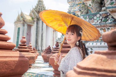 Geleneksel Tayland elbisesi giyen genç bir kadın, dünyanın dört bir yanındaki turistler için popüler bir yer olan Wat Arun 'da antik bir şemsiye tutuyor. Bangkok Tayland