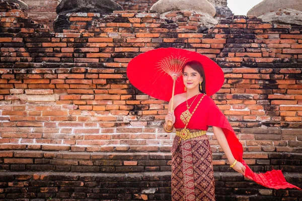 伝統的な赤いタイのドレスと黄金のアクセサリーを身に着けている若い女性の閉鎖は 歴史的なサイトで伝統的な傘を持って立っています タイ民族衣装 — ストック写真