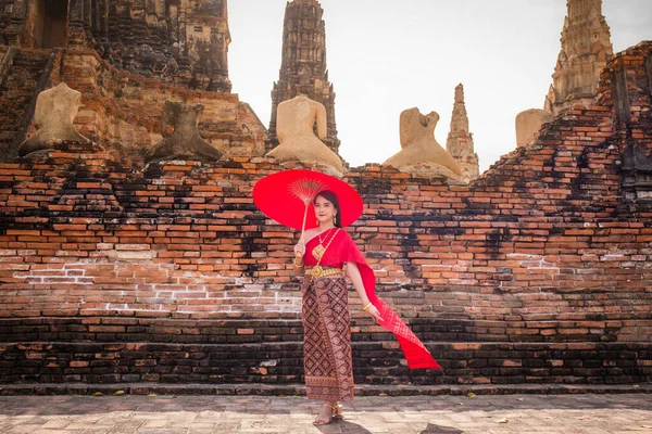 伝統的な赤いタイのドレスと黄金のアクセサリーを身に着けている若い女性は 歴史的なサイトで伝統的な傘を持って立っている タイ民族衣装 — ストック写真