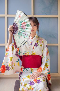 Japon tarzı kimono giyen, elinde kağıt yelpaze tutan genç bir kadın Japon tarzı bir evde oturuyor.