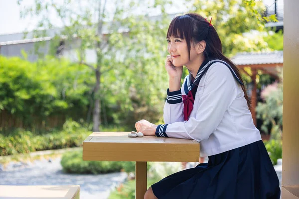 日本の学校の制服を着た女性の閉鎖は 多くの緑の木がある公園で伝統的な木製の椅子に座っています — ストック写真