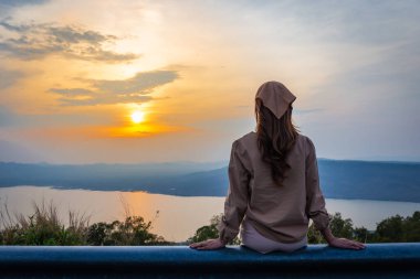 Kadın arkasına yaslanmış, arkasında dağlar ve nehirle gün batımına bakıyor.