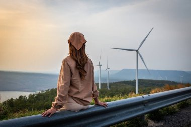 Kadın gün batımını seyrediyor. Arkasında dağlar, nehir ve rüzgar türbini var.