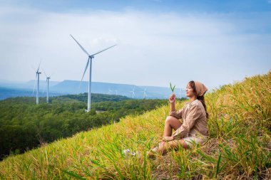 Hoş bir elbise giymiş bir kadın arka planda dağlar ve rüzgar türbini olan bir çayırda oturuyor. Seyahat özgürlüğü kavramı