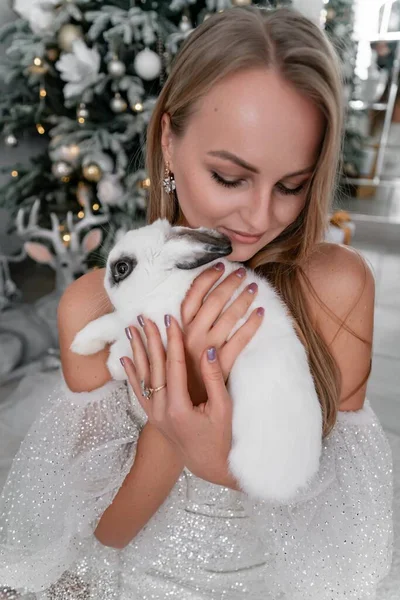 2023年の白ウサギのシンボルを持っている女性 輝かしいドレスでウサギを抱えている美しい若い金髪の女性のクローズアップ 彼女はクリスマスの装飾された部屋に座っている — ストック写真