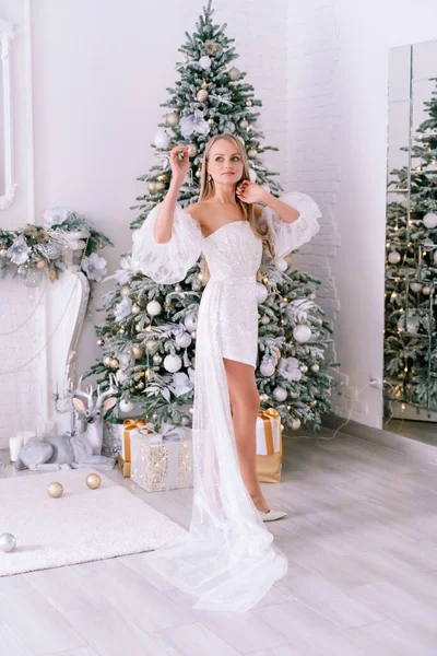 クリスマスルームのブロンド 光沢のある明るい短いドレスの美しいブロンドの女性は クリスマスツリーでお祝いのインテリアで装飾された美しい明るい部屋に立っています — ストック写真