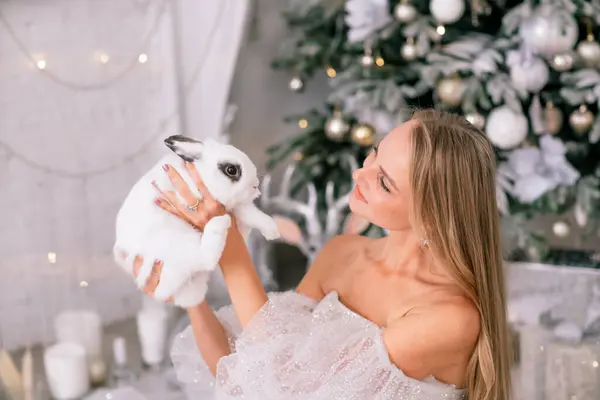 2023年の白ウサギのシンボルを持っている女性 輝かしいドレスでウサギを抱えている美しい若い金髪の女性のクローズアップ 彼女はクリスマスの装飾された部屋に座っている — ストック写真