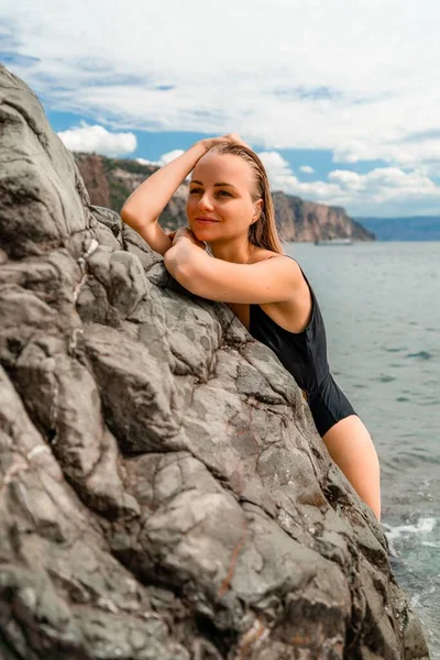 水着の海を泳ぐ女性 黒い水着の魅力的なブロンドの女性は 岩の周りの海岸の空気を楽しんでいます 旅行と休暇のコンセプト — ストック写真