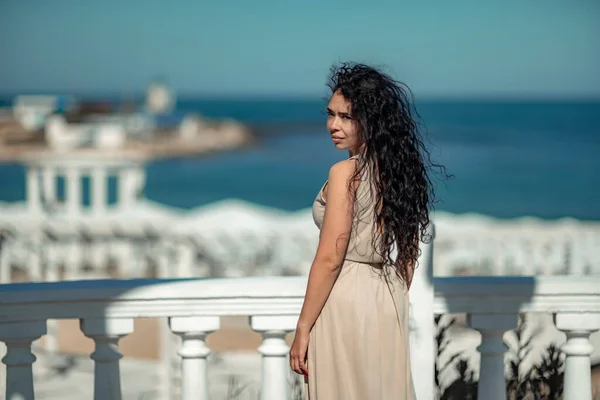 Sea woman rest. A woman with long curly hair in a beige dress stands with her back and looks at the sea and the coast from a balcony with balusters. Tourist trip to the sea