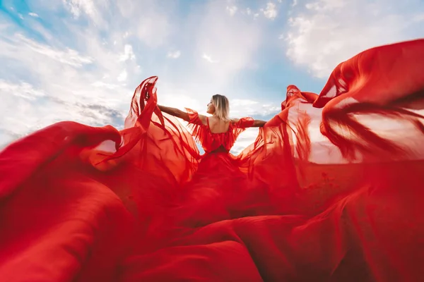 Woman Red Flying Dress. A blonde in a red dress against the sky. Rear view of a beautiful blonde woman in a red dress fluttering in the wind against a blue sky and clouds
