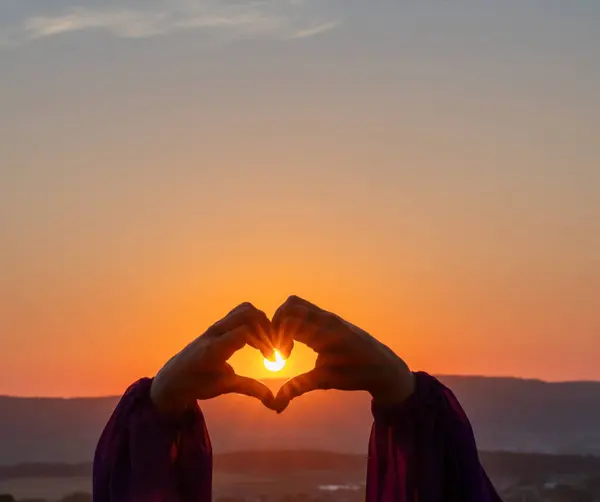 woman creates form a heart shape with hands at sunset. Conveying serene sunset ambiance with heart gesture