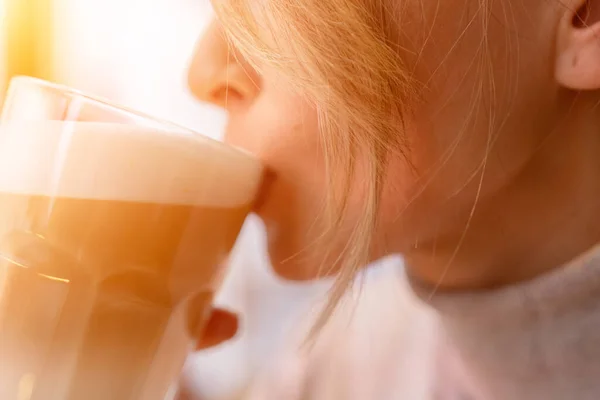 Woman with blonde hair sips cappuccino in a cafe. She is holding the glass up to her face, taking a sip of the drink