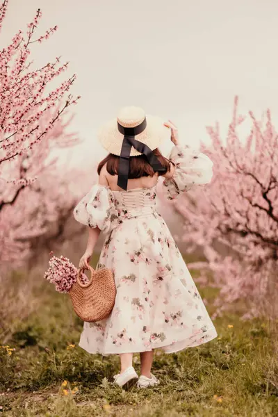 Woman blooming peach orchard. Against the backdrop of a picturesque peach orchard, a woman in a long dress and hat enjoys a peaceful walk in the park, surrounded by the beauty of nature
