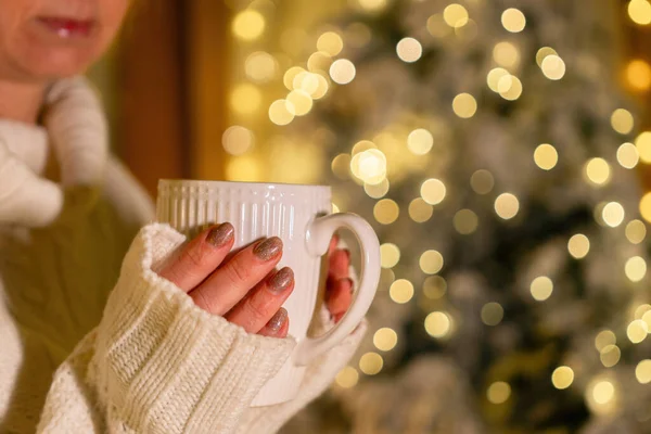 White mug in hands on a magical bokeh background. Beautiful Christmas gift boxes with ribbon.