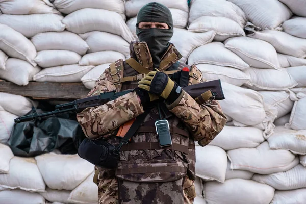 stock image Soldier with weapon in military uniform stands next to the barricades made from sandbags and anti-tank hedgehog barriers. Military man on the roadblock. Combatant in full ammunition. Concept of war