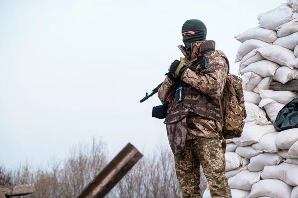 stock image Soldier with weapon in military uniform stands next to the barricades made from sandbags and anti-tank hedgehog barriers. Military man on the roadblock. Combatant in full ammunition. Concept of war