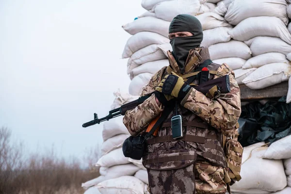 stock image Soldier with weapon in military uniform stands next to the barricades made from sandbags and anti-tank hedgehog barriers. Military man on the roadblock. Combatant in full ammunition. Concept of war