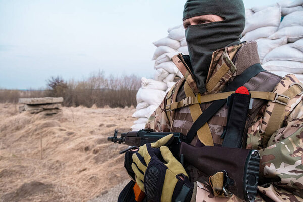 Soldier with weapon in military uniform stands next to the barricades made from sandbags and anti-tank hedgehog barriers. Military man on the roadblock. Combatant in full ammunition. Concept of war