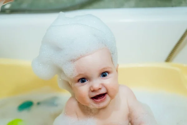 Baby girl in a bath with foam and soap bubbles. Happy laughing baby taking a bath playing with toys. Little child in a bathtub. Smiling kid in bathroom. Infant washing and bathing