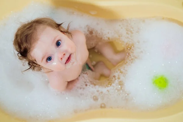 Baby girl in a bath with foam and soap bubbles. Happy laughing baby taking a bath playing with toys. Little child in a bathtub. Smiling kid in bathroom. Infant washing and bathing