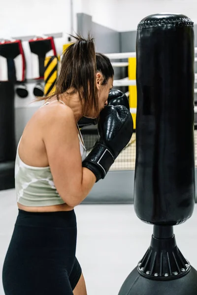 Vertical image of a young woman with black boxing gloves exercising with a punching bag in gym.
