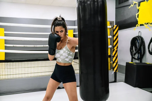 Front view of a confident young woman with black boxing gloves punching bag in gym.