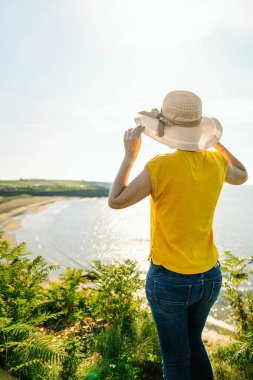 Renkli sarı tişörtlü, tanınmaz haldeki turist kız şapkasına dokunuyor, öğlen güneşli yeşil giysili uçurumdan sahile bakıyor..