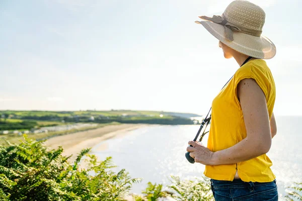 stock image A casual, blonde tourist wearing a colorful yellow tee and a hat, capturing a coastal landscape with lush green vegetation from a cliff, under the bright midday sun.