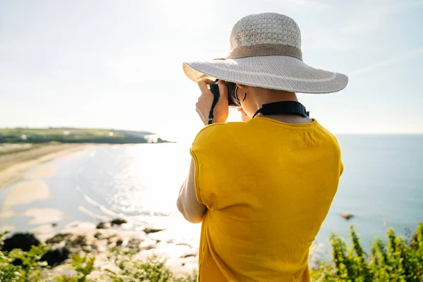 stock image A casual blonde tourist in a colorful yellow tee and hat captures a coastal landscape from a lush green cliff with bright noon sunlight.