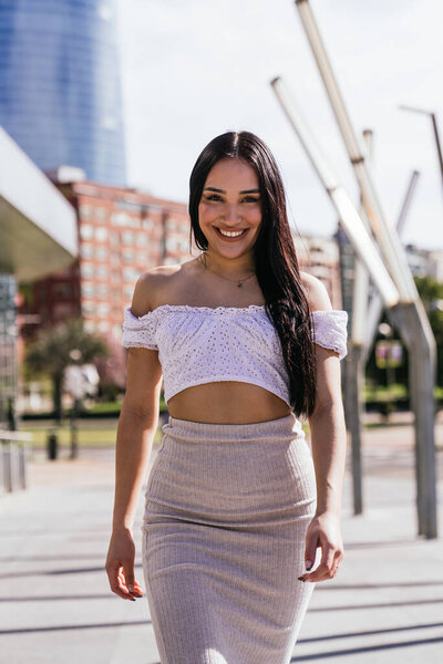 This street portrait captures a beautiful and cheerful Latina girl wearing casual and modern clothes, smiling on a sunny day in the middle of the street