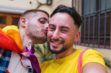 LGBTQ aksesuarlarıyla süslenmiş iki neşeli eşcinsel arkadaş, canlı evlerini terk ederken bir selfie ve bir öpücük paylaşırlar..