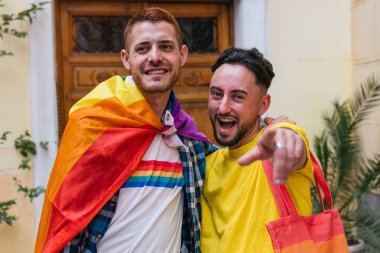 Mutluluk saçan iki eşcinsel arkadaş, sıkıca sarılıyorlar. LGBT ürünlerini gururla sergiliyorlar, evlerinden dışarı adım atıyorlar, kameraya neşeyle bakıyorlar..