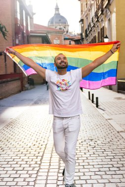 Sakallı yakışıklı bir adam gururla bir LGBT bayrağı tutuyor, güneşli bir şehir sokağında, gün batımında gökyüzüne bakıyor..