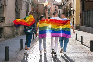 Alacakaranlıktaki güneşli şehir manzarası, LGBTQ sembolleriyle gururla süslenmiş, kol kola mutlu bir şekilde yürüyen, birlik ve gurur gösteren dört eşcinsel arkadaş için zemin görevi görüyor..