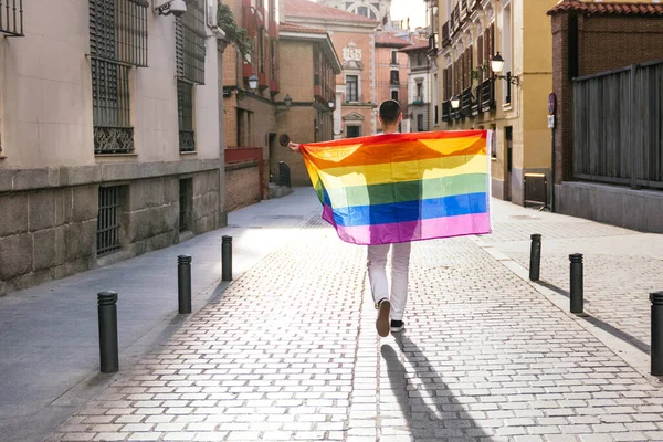 stock image A man holds the LGBT flag, his back to the camera, arms open wide, basking in the sunlit city street during a vibrant sunset.