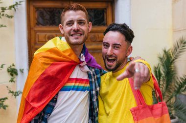 Mutluluk saçan iki eşcinsel arkadaş, sıkıca sarılıyorlar. LGBT ürünlerini gururla sergiliyorlar, evlerinden dışarı adım atıyorlar, kameraya neşeyle bakıyorlar..