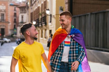 Batan güneş tarafından aydınlatılmış, el ele yürüyen iki eşcinsel arkadaşın yakın plan fotoğrafı. Canlı LGBTQ aksesuarları onları süslüyor..