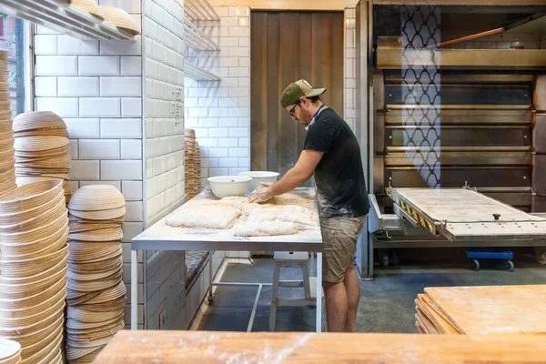 一个年轻人在面包店里巧妙地把小麦面团做成面包条 面包烘烤的热烘烘炉旁边放满了面粉的混合物 — 图库照片