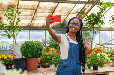 Coşkulu bir Afro-Amerikan işçi renkli bir çiçeklikte bir selfie çekiyor, kamerayı canlı bir bitki ve çiçek zeminiyle selamlıyor.. 