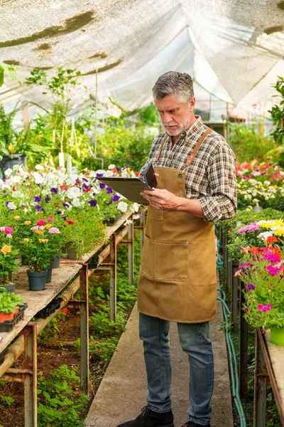 A 55-year-old worker in a vivid plant nursery, documenting his tasks and customer inquiries in a notebook.