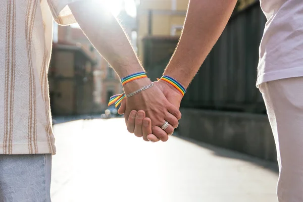 stock image Close-up photo of two unidentified men's hands, tightly holding each other while walking along a sun-drenched city street at sunset. They wear rainbow-colored LGBT bracelets on their wrists.