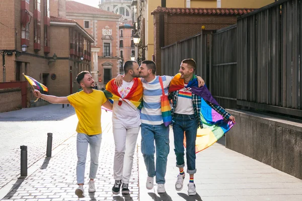 stock image In a sun-kissed city street, four exuberant gay friends, adorned with LGBT pride accessories, joyfully stroll amidst the sunset's glow. Two share a kiss.