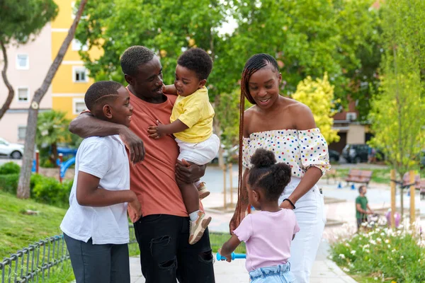 Çağlar ve cinsiyetlerden oluşan ışıldayan Afrikalı bir aile, kucağında bebek olan bir baba, canlı bir şehir parkında sevgi dolu bir kucaklaşmayı paylaşıyor..