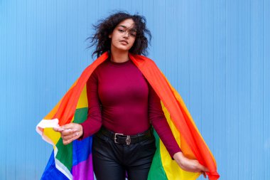 Siyah saçlı beyaz bir kadının büyüleyici fotoğrafı mavi bir arkaplanda canlı bir LGBTQ + bayrağı sergiliyor.