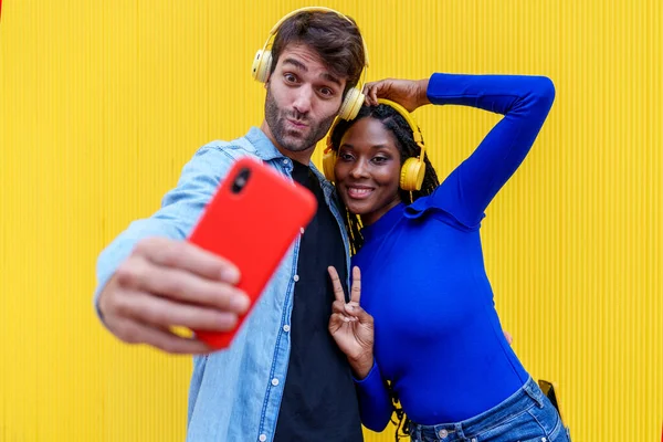 Stock image Vibrant selfie of two friends, a Caucasian man and an African woman, immersed in music, sharing laughter and joy against a yellow background