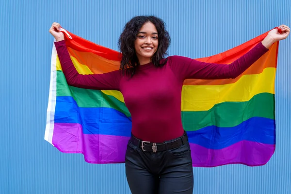 A vibrant, colorful photograph of a happy, black-haired Caucasian woman proudly holding an LGBT rainbow flag against a bright blue background