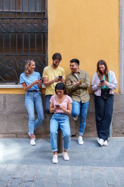 Gençlik Farklılığı: Canlı şehir kıyafetleri içinde beş arkadaş, bir şehir binasına yaslanmış, telefonlarına dalmış, çevrelerinden habersiz.
